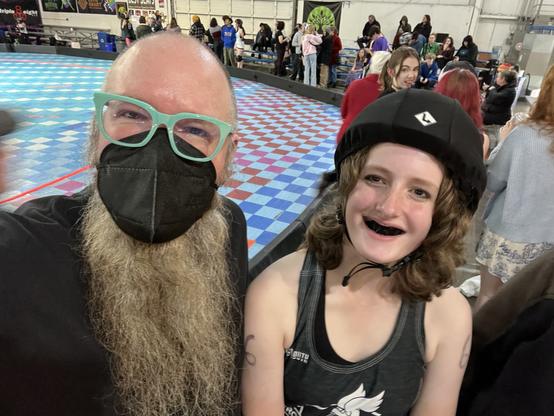 Me with my long beard, sea foam glasses, and mask. I’m standing next to Ro with her black kit and black mouthguard. We’re smiling with a colorful checkered rink behind us.