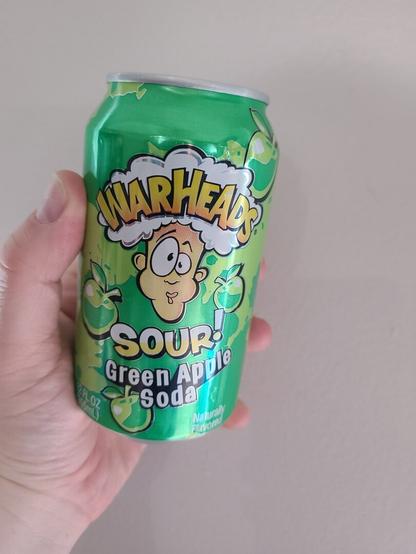 White person's hand holding a green soda can against a light colored wall. The can is labeled "Warheads Sour! Green Apple Soda." The center of the can has the Warheads logo, which is a cartoon face puckering their lips extremely tight as though they are eating something sour. Cartoonish green apples are strewn across the can in various locations behind the text / logo.