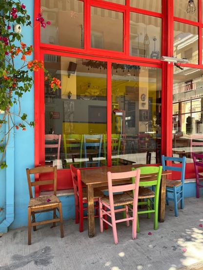 There’s a blue wall and a window of large panes of glass with aluminium frames painted red and a wooden table with straw-seated wooden chairs, wood stained and painted - pink, green blue. Inside another row of painted chairs. It’s a taverna, closed.