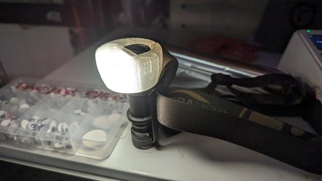 A zebralight headlamp, magnetically mounted on a metal surface. On top of the torch, is a 3d printed cap that diffuses the light into a wider area, so the headlamp can act as a table light.