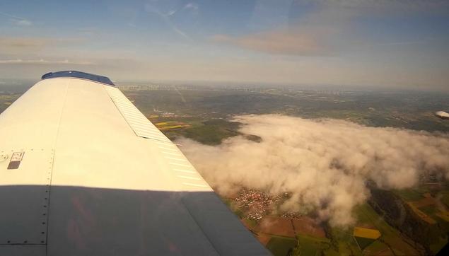 Blick aus dem Fenster auf die rechte Tragfläche eine Kleinflugzeugs, blauer Himmel mit wenigen Wolken; unten Felder und Dörfer mit tiefen Wolken/Bodennebel.