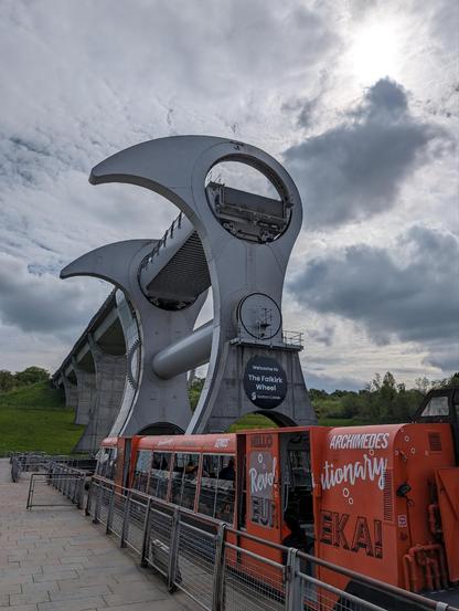 Dockside on a cloudy day with a canal barge boat nearby. In the background there is an engineering structure best described as metal bottle openers or stylistic birds with beaks pointing left, connected by cylindrical tubes. They can lift the boat and rotate it around while another is brought down.