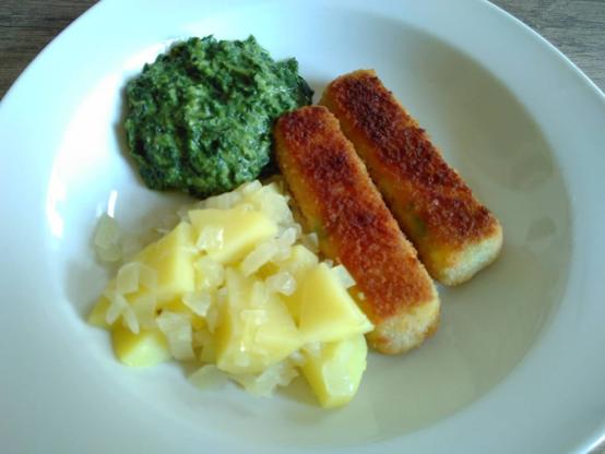 Ein weißer Teller mit zwei gebräunten Gemüsestäbchen rechts (im Aussehen wie Fischstäbchen), links hinten veganer Rahmspinat, davor gestückelte Kartoffeln mit Zwiebelstückchen.