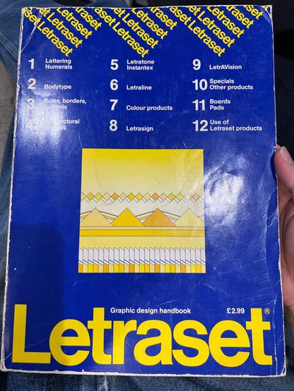 The cover of a Letraset graphic design handbook. It’s blue with a large Letraset logo in yellow at the bottom, with ‘Graphic design handbook’ and ‘£2.99’ above it. There is a square image in the middle which is a series of squares, triangles and boxes in various shades of yellow.  At the top are some small, repeated Letraset logos at roughly 45° in yellow with a list of the sections features in the catalogue below them.