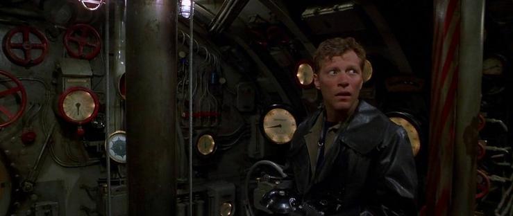 A young man in a leather jacket stands inside an old submarine, looking around