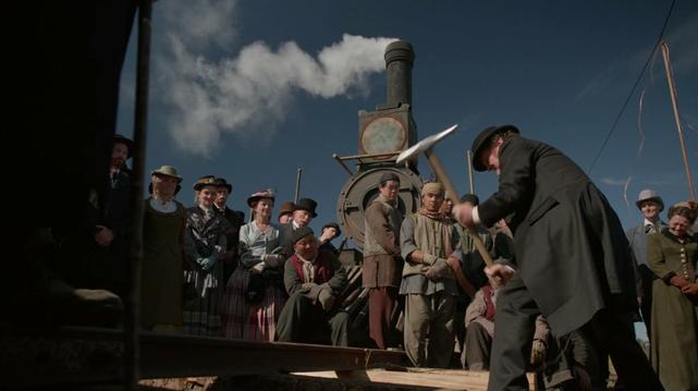 A scene from an old-fashioned movie. It shows a white man pounding in a final railway spike as people look on