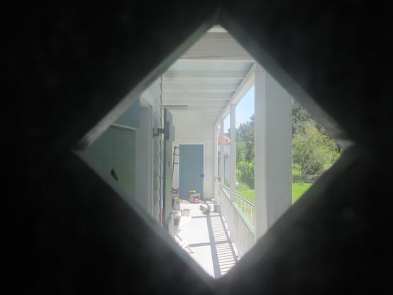 View out through square window set with points at top bottom and sides, diamond shape.  Through window is a long narrow roofed porch of a wooden building. Along the left side and in distance straight ahead are wooden shutters covering windows and doors. On the porch are various small items, including cans of paint, a bucket, small folding tables, a leaf blower, and a bag of charcoal.
At right, stairs lead down about a meter to the grounds.   Greenery of lawn, small trees and large leafed plants.