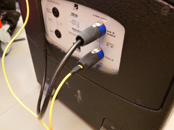The USB2Speakon plugged into the back of a subwoofer with pass-through and a connected yellow USB-C cable leaving the image.
