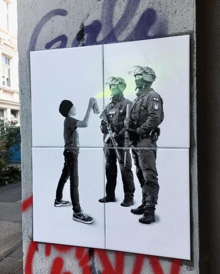 Streetartwall. Four white, painted tiles are attached to a gray house wall as street art. The picture is mainly in black and white and shows two police officers with helmets, visors and machine guns. They are standing opposite a boy in jeans, a T-shirt, sneakers and a woolly hat, who is spraying green paint on their faces with a spray can.
Info: The Artist "Adultremix" creates its socio-cultural, critical collages with a worldwide unique stencil technique and laserwriter toner. The manipulation of the printer results in a new art technique. The works are also available in print.