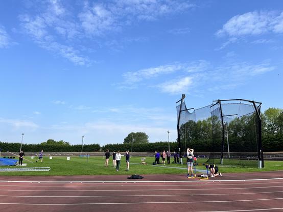 Outdoor athletics stadium with discus cage in foreground, athletes waiting. And glorious blue sky!