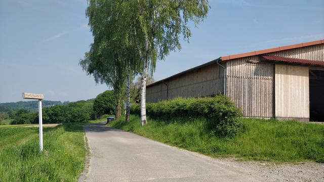 Einfahrt zur Kornkammer Holte. Neben einem Feld steht ein Holzschild "Ruhrtalgarten". Rechts ist eine Lagerhalle. Die Kornkammer liegt auf einem Hügel, sodass man das Ruhrtal überblicken kann.