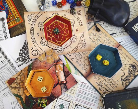Foto de tres bandejas lanzadados con diseños distintos. Una como la mesa de un mago, otra como un cuenco de bronce en la mesa de una taberna, y otra como un diario lovecraftiano.