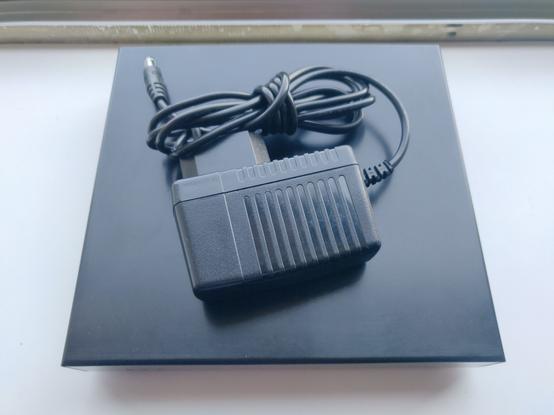 Pcengines APU4D4 im schwarz eloxiertem Gehäuse, auf dem das dazu gehörige Stecker Netzteil liegt.