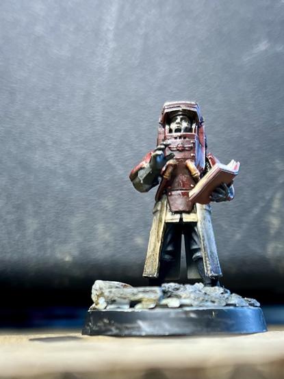 A priestly figure in a mechanical outfit reads aloud from a red book