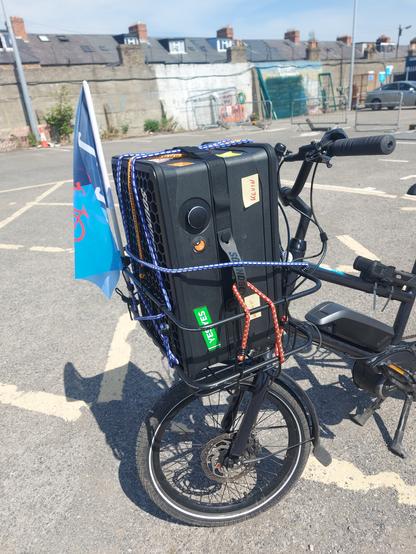 Black Orbea Katu small wheeled bike carry Soundboks speaker in front basket secured with bungee strap and parking in near empty carpark at Dalymount Park.