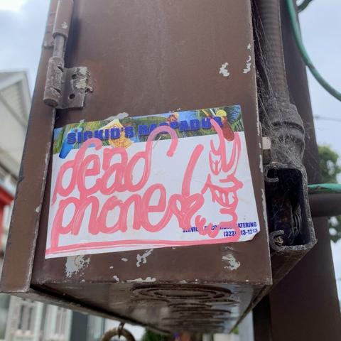 A slap sticker on a post. It says "Dead Phone" in a nice cursive style and has a drawing of a person's face as they smirk towards the viewer. The art is done with a pink paint pen on a sticker label that says "Sickid's Respados" on the top.