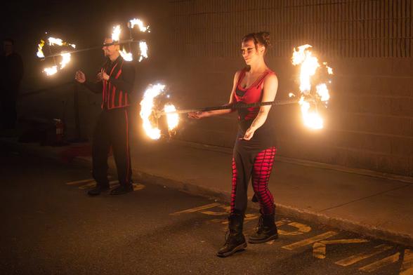 Two performers are doing a fire show at night, with the woman and man spinning fire staffs. They are wearing performance costumes.