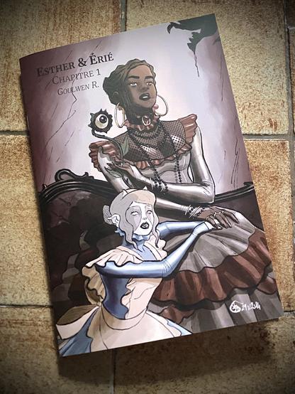 Couverture du 1er Chapitre d’Esther et Erié

Esther est assise sur un canapé de style victoirien, elle porte une robe d’époque en soie grise, avec volants rouges et de nombreux bijoux et des boucles d’oreilles créoles. Dans sa main gauche, elle tient une fleur étrange avec un œil à la place du pistil.

Erié est à genoux à ses pieds, dans sa tenue de servante. Ses mains sont sous la main droite d’Esther, posées sur les genoux de la maîtresse.

Les deux semblent regarder la créature qu’on aperçoit dans le coin supérieur droit, Esther comme hypnotis, tandis qu’Érié semble inquiète