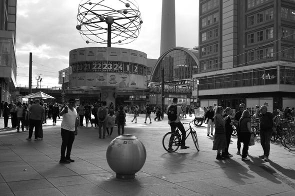 Black and White photo taken at Berlin Alexanderplatz. In front is the large (famous) world time clock. In the back you see Alexanderplatz station. To the right of the image there's a man taking a selfie with the clock.