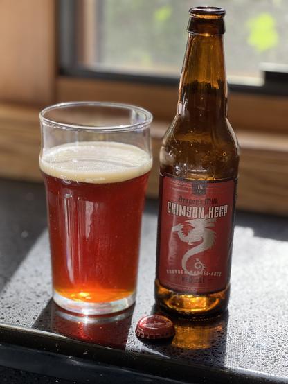 A pint glass of ruby red ale, with the bottle to the left. The label is deep red with a silver dragon with unfurled wings in silhouette