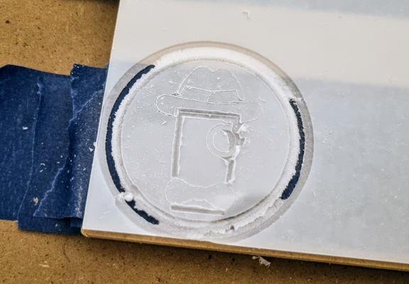 A CNC router workpiece stuck down to the cutting bed, it's a sheet of perspex with opaque white protective film on it, a logo is carved into it with a very rough cut out circle around it.
