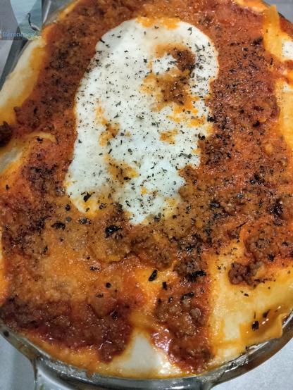 A vertically-oriented photograph of the top of a pan of lasagna. The pan is oval-shaped. There is a white oval in the inside, surrounded by red meat sauce, surrounded by fresh pasta.