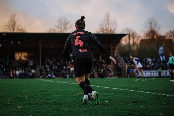 Sylwia Matysik am Ball, mit dem Rücken zur Kamera. Sie trägt das schwarze Auswärtstrikot Leverkusens mit der hellroten Rückennummer 4 und ihrem Namen darauf. Der Himmel über ihr ist von der Abenddämmerung blau-orange. Ihre Silhoutte und die Tribüne zeichnen sich gegen den Himmel ab, Matysik nimmt gerade Anlauf, um einen Freistoß zu treten.