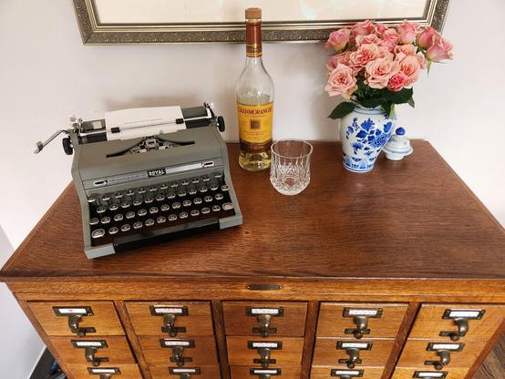A wooden library card catalog on which sits a 1949 Royal Quiet De Luxe Typewriter next to a crystal old fashioned glass and a fifth of Glenmorangie in a scotch bottle. To one side is a blue vase with small pink roses.