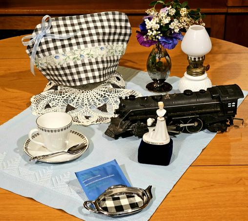 A tea tableau of an antique aqua blue tea towel atop which is a Harvey Girl figurine standing beside a heavy black metal train engine, accompanied by a milk glass hobnail kerosene lantern, vintage china, and a mercury glass vase of flowers. The tea cozy is black & white gingham with a pale blue gauze bow, & light green & blue calico ribbon trim. There is a black & white glass- beaded doily under the teapot.