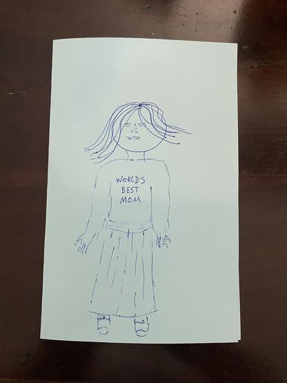 Poorly drawn stick figure of my mom wearing a “Worlds Best Mom” shirt on the front of my handdrawn card.