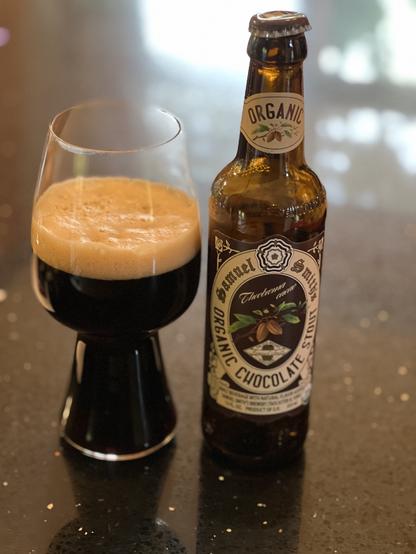 A stout glass of dark beer next to a brown bottle with a white and brown label. Central to the label are cocoa beans and green leaves.