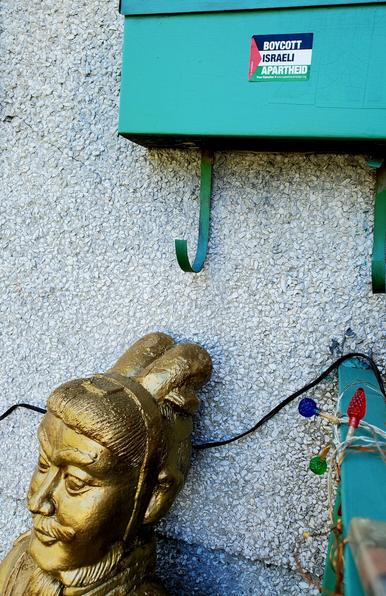 Front mailbox with a BOYCOTT ISRAEL APARTHEID sticker on right & gold Chinese guard statue to left. Coloured outdoor light string is running through.