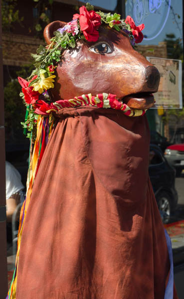 A brown fiberglass bear head, wearing a garland of red roses, with a red and yellow ruffle under her chin, and a body consisting of a cloak of thick brown fabric