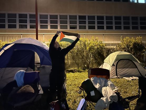 A student in front of tents holding up a Palestinian flag, light glowing through it.