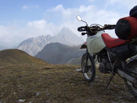 Auf einem Grasbedeckten Bergrücken ragt eine mit Packsack beladene Enduro von rechts ins Bild. Im Hintergrund hinter leichtem Dunst eine felsige, schroffe Bergspitze.