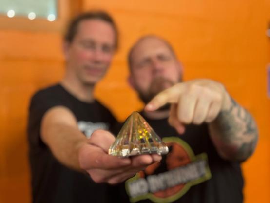 Nicolas und Reinhard stehen vor einer orangenen Wand, der Hintergrund ist verschwommen. Nicolas hält in der Hand eine Schwurbel-Pyramide, die Reinhard mit der Fingerspitze berührt.