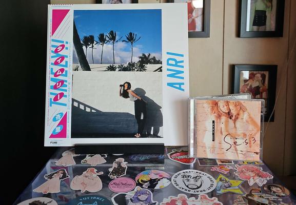ANRI - TIMELY!! vinyl album is displayed on top of a turntable plastic cover. To the right is S.E.S - LOVE on CD. The turntable cover is covered in various stickers, and on the wall behind are some framed pictures. 

ANRI's album cover shows ANRI standing in front of a wall looking off in the distance. S.E.S album cover shows the 3 members standing close together.