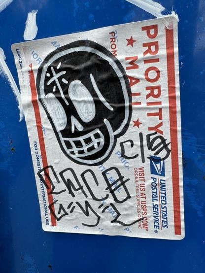 A USPS label that has graffiti art on it and is stuck on a wall. The art shows a large black skull with white lines and text that says "Taco C/S". The skull is in the style of Mexican folk art and the text is written in Chino style graffiti handwriting.