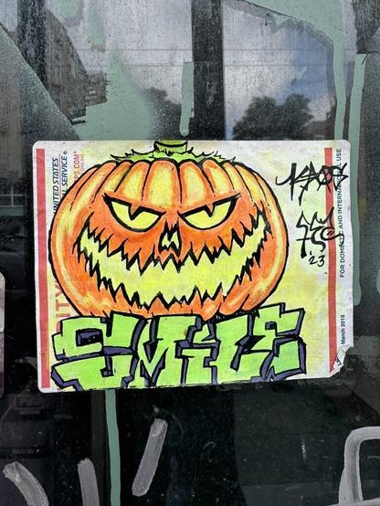 A USPS sticker with a drawing of a Jack O Lantern on it. The art is drawn in sharpie and coloring pencil. In big graffiti style letters, the artist wrote "Smile". They also wrote "Kaos" in a smaller handwriting, which I think is their signature. This sticker is stuck to a window.