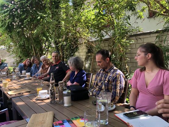Ein Tisch in einem Garten, daran sitzen viele Menschen und unterhalten sich. In der Mitte Katrin, von der Seite.