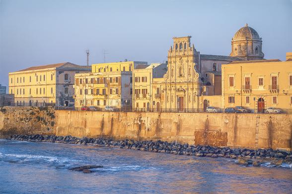 Golden early morning light bathes the picturesque historic buildings along the waterfront in Ortigia, Sicily, Italy. A seawall separates the waterfront building from the rocky shore and the sea.