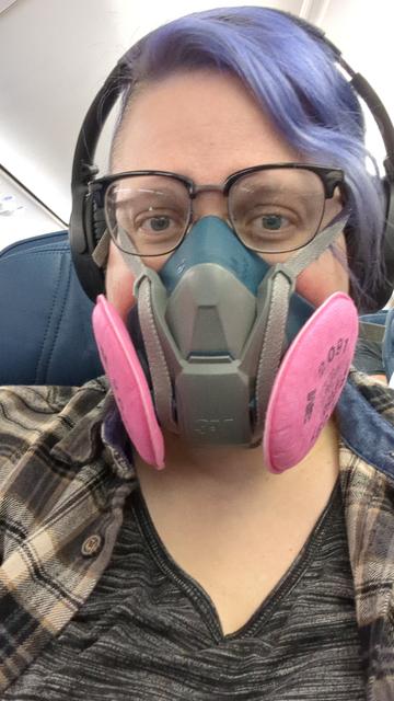Person with purple wavy hair in a brown flannel shirt wearing glasses and a N95 painter mask.