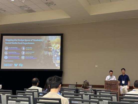 Two speakers in front of a podium next to a big screen with a title slide