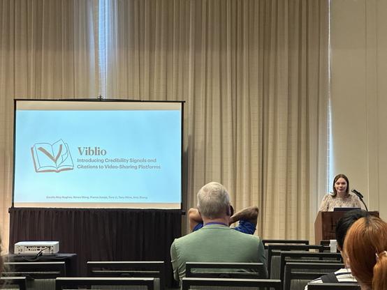 A speaker behind a lecturn next to a big screen with a title slide