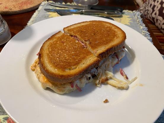 Photograph of a grilled sandwich on a white plate, with melted cheese, sauerkraut, and bits of meat spilling out.