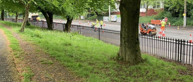 Beyond the trees, workmen are pouring tarmac. Beside a small road roller, a man with a stop sign directs traffic. The new segregated cycle lane has a pristine surface.