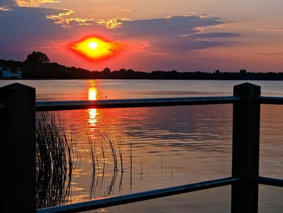 The sun is setting over the lake and the forest on the other shore. The yellow sun is surrounded by a red aura and yes, it reminds me of the Eye of Sauron, too 😆 The light of the sun is reflected in the water. The railing of a jetty can be seen in the foreground of the picture.