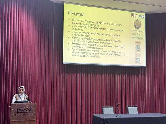 A speaker in front of a lectern on a stage with a big screen with takeaways on a slide