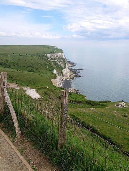 Blue sky with wispy white clouds above white cliffs in the middle of the shot dropping down to a black pebble beach and sea for the whole right half of the shot. In the foreground, grassy slopes leading down to the sea with a fence visible with wooden fence posts.