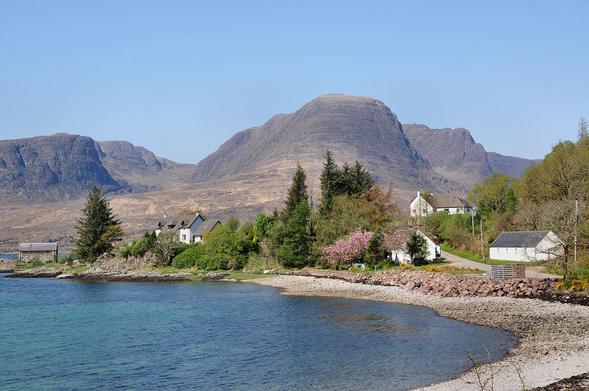 Kishorn in Wester Ross. The image shows a view over a bay with a stony beach that faces left out to sea. There’s a headland beyond the bay on which are trees and houses. In the background is a glimpse of more water, and then in the distance there are mountains with dark rocky faces rising to considerable height. The sky is blue.